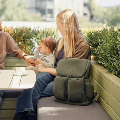 Woman with a green backpack sitting on a bench outdoors, with a child and another person in the background.
