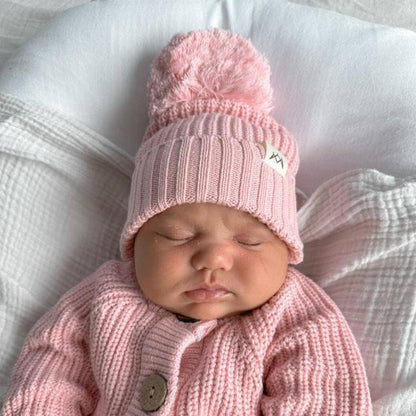Newborn baby wearing a pink knitted outfit with a matching hat on a soft surface.