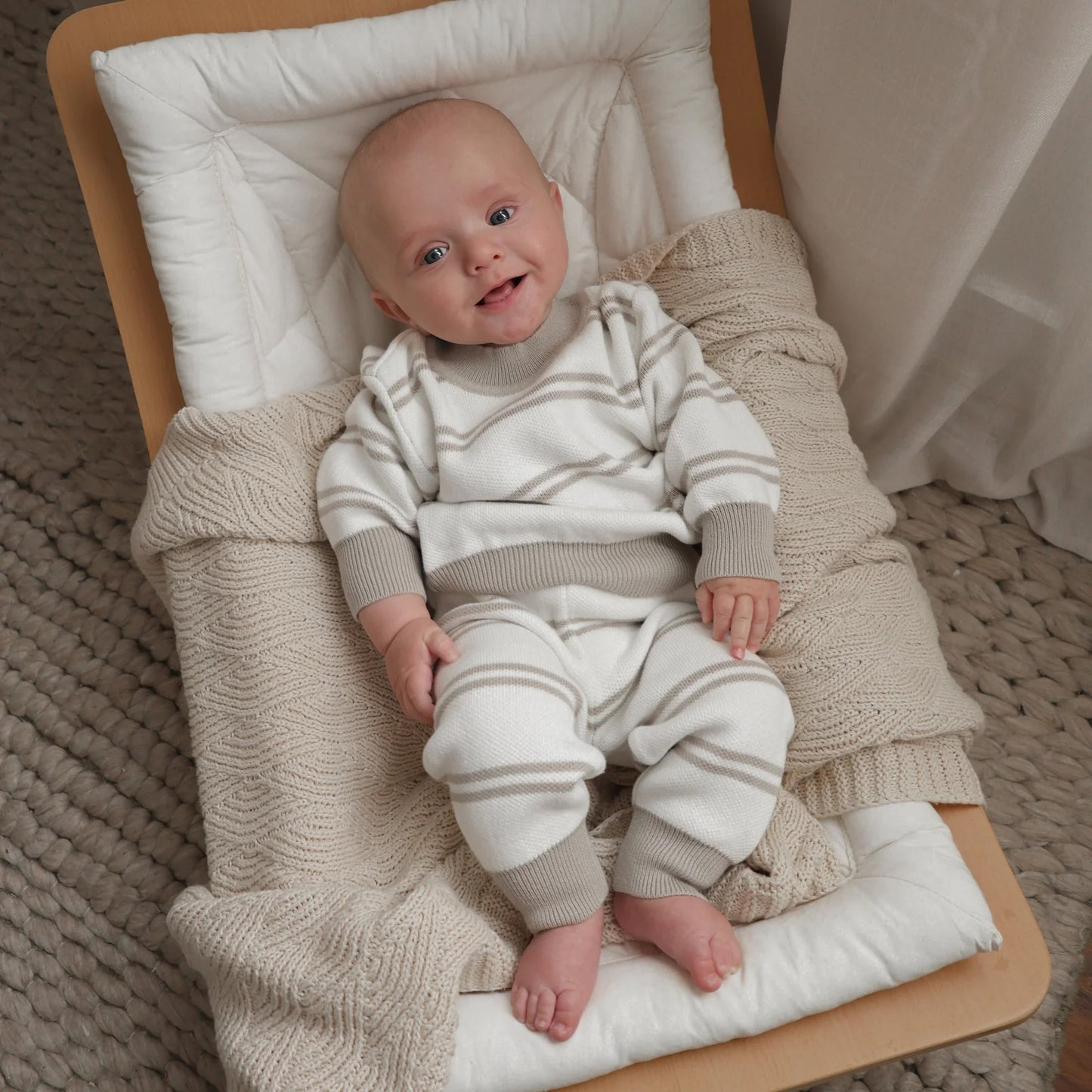 Baby in a striped outfit sitting on a wooden crib with a beige blanket