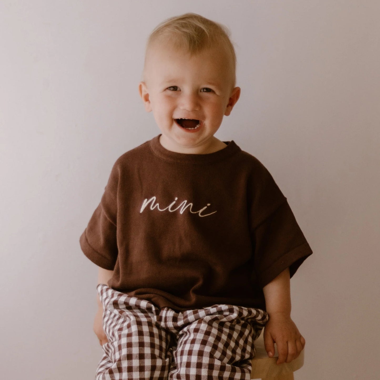 Child wearing a brown shirt with 'mini' text, sitting on a wooden stool against a plain background