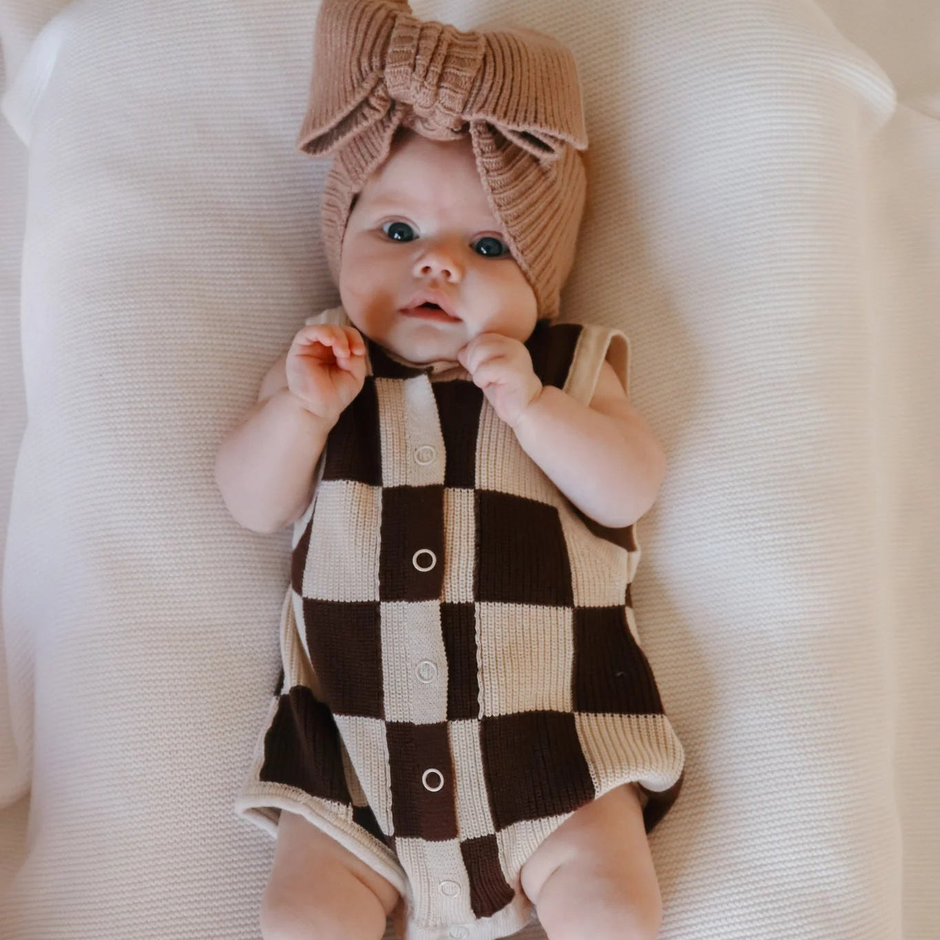 Baby wearing a checkered romper and headband on a white surface