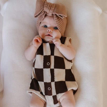 Baby wearing a checkered romper and headband on a white surface