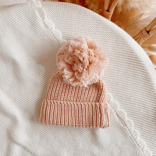 Pink knitted beanie with a fluffy pom-pom on a textured surface