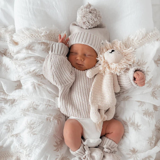 Newborn baby in a cozy outfit with a knit hat and scarf, lying on a textured blanket.