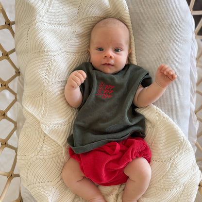 Baby wearing a dark shirt with red text and red shorts sitting on a textured beige surface.