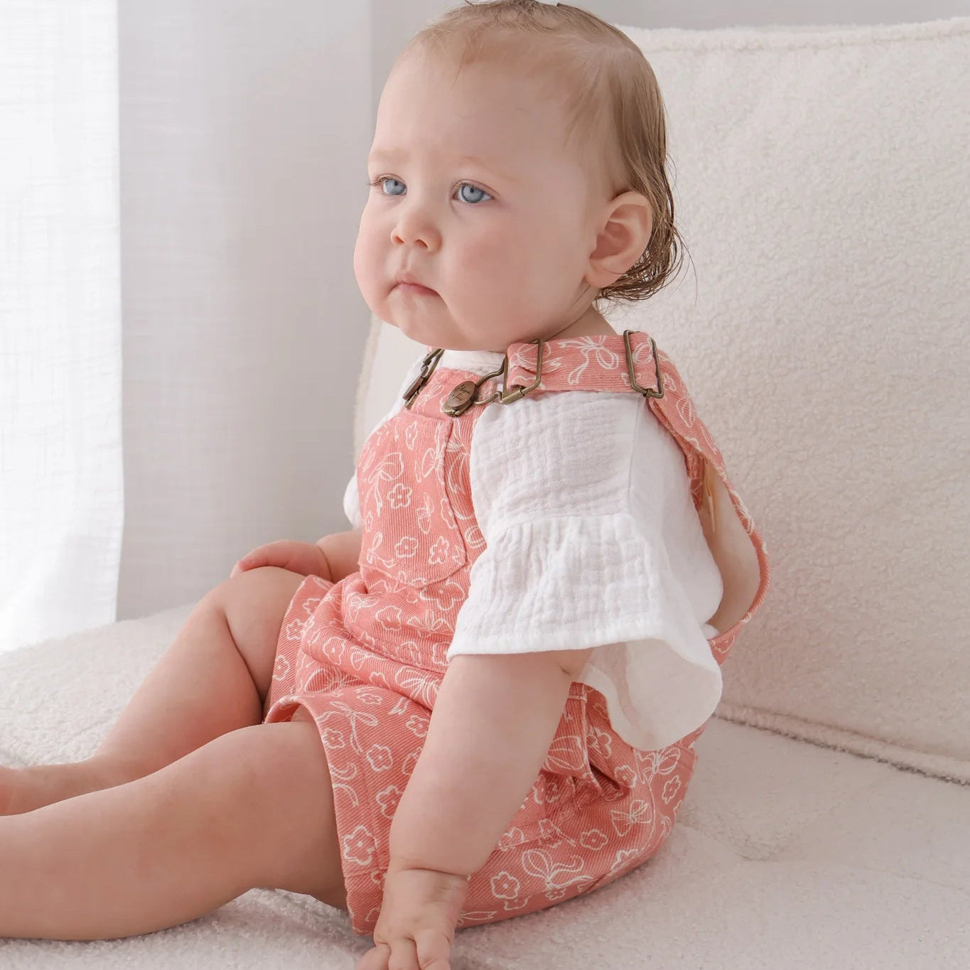 Baby sitting on a couch wearing a pink patterned dress and white shirt.