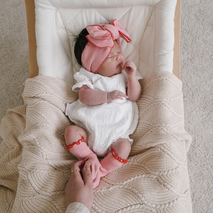 Newborn baby in a crib with a pink bow and white outfit, surrounded by soft textures.