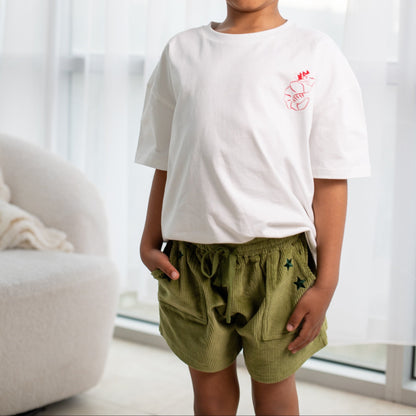 Child wearing a white t-shirt with a red logo and green shorts standing in a room with white curtains.