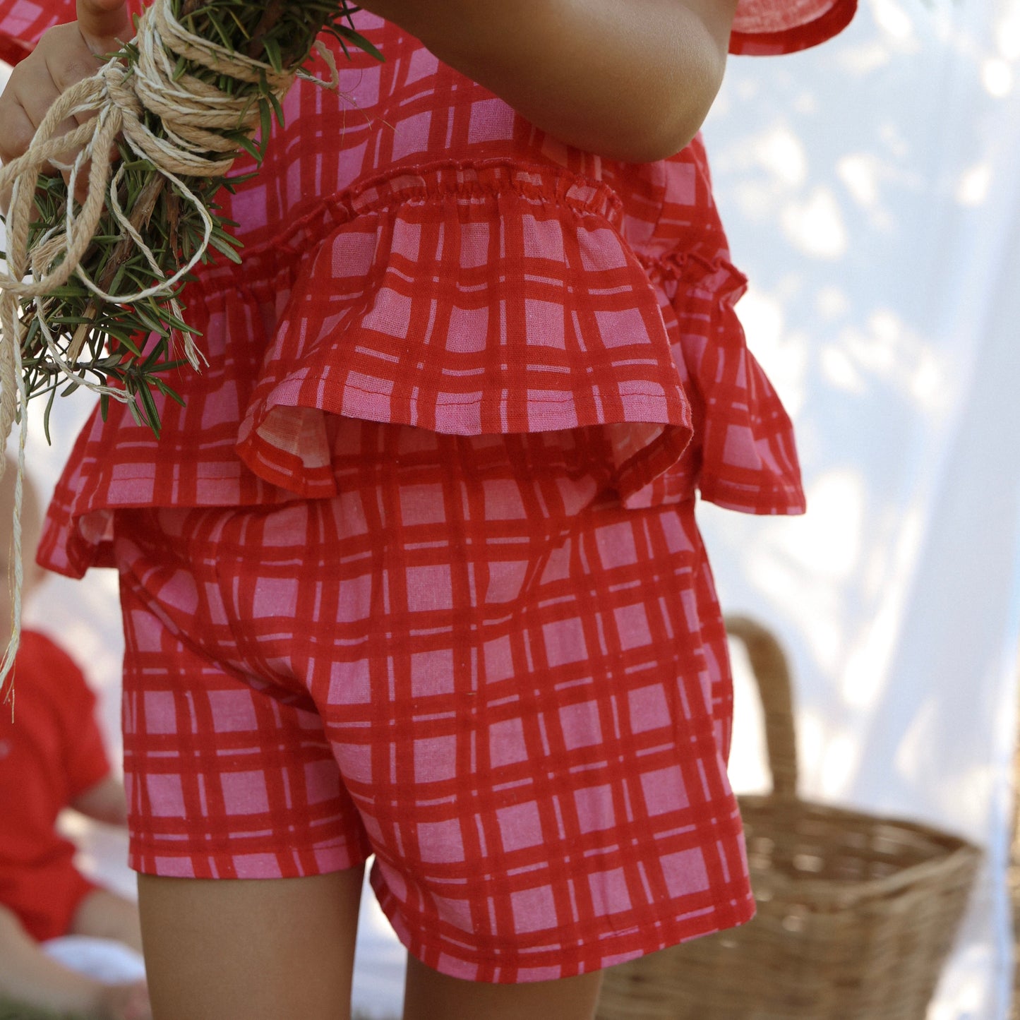 Child wearing a red and white checkered outfit holding a bundle of greenery.