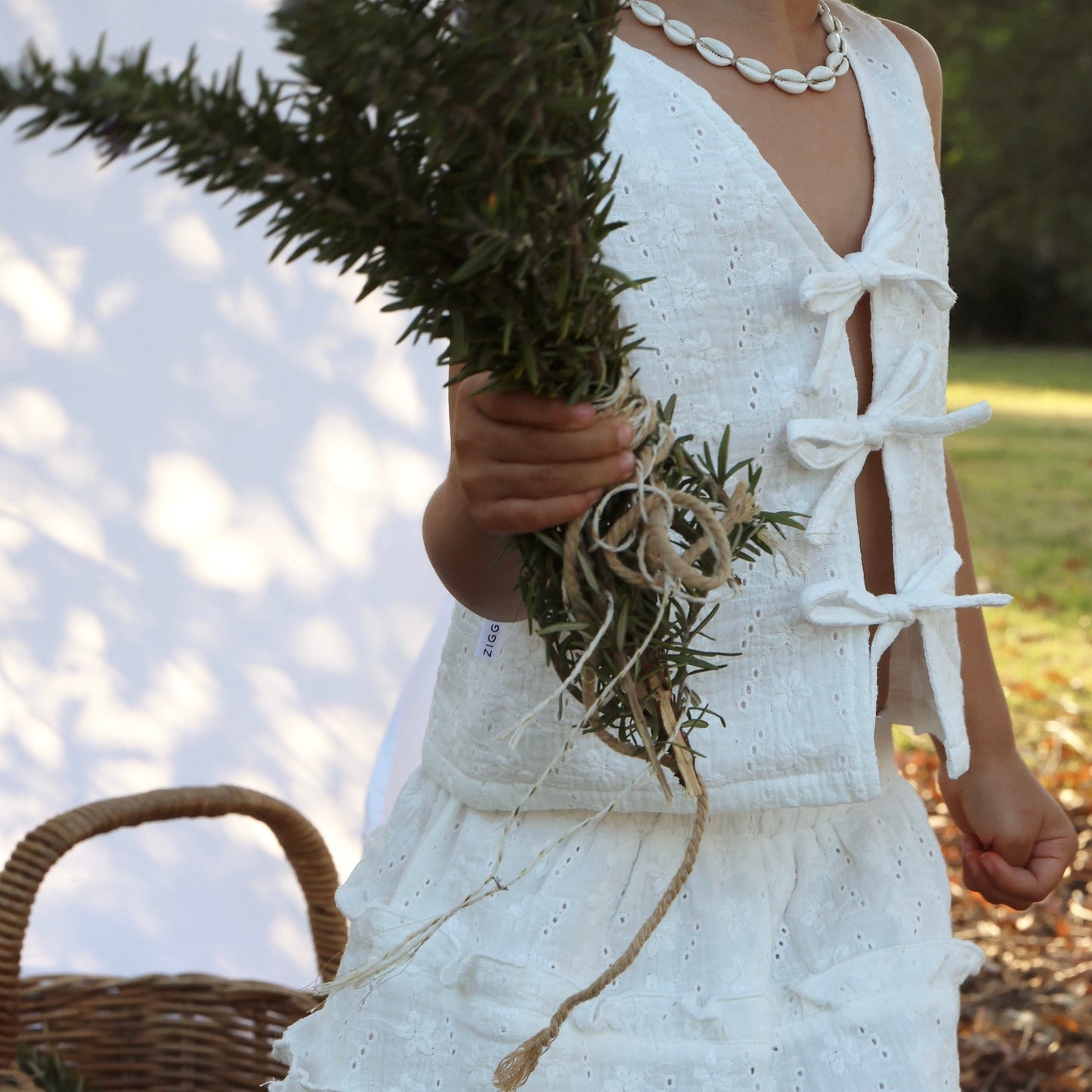 Person in a white dress holding greenery with a wicker basket on a natural background