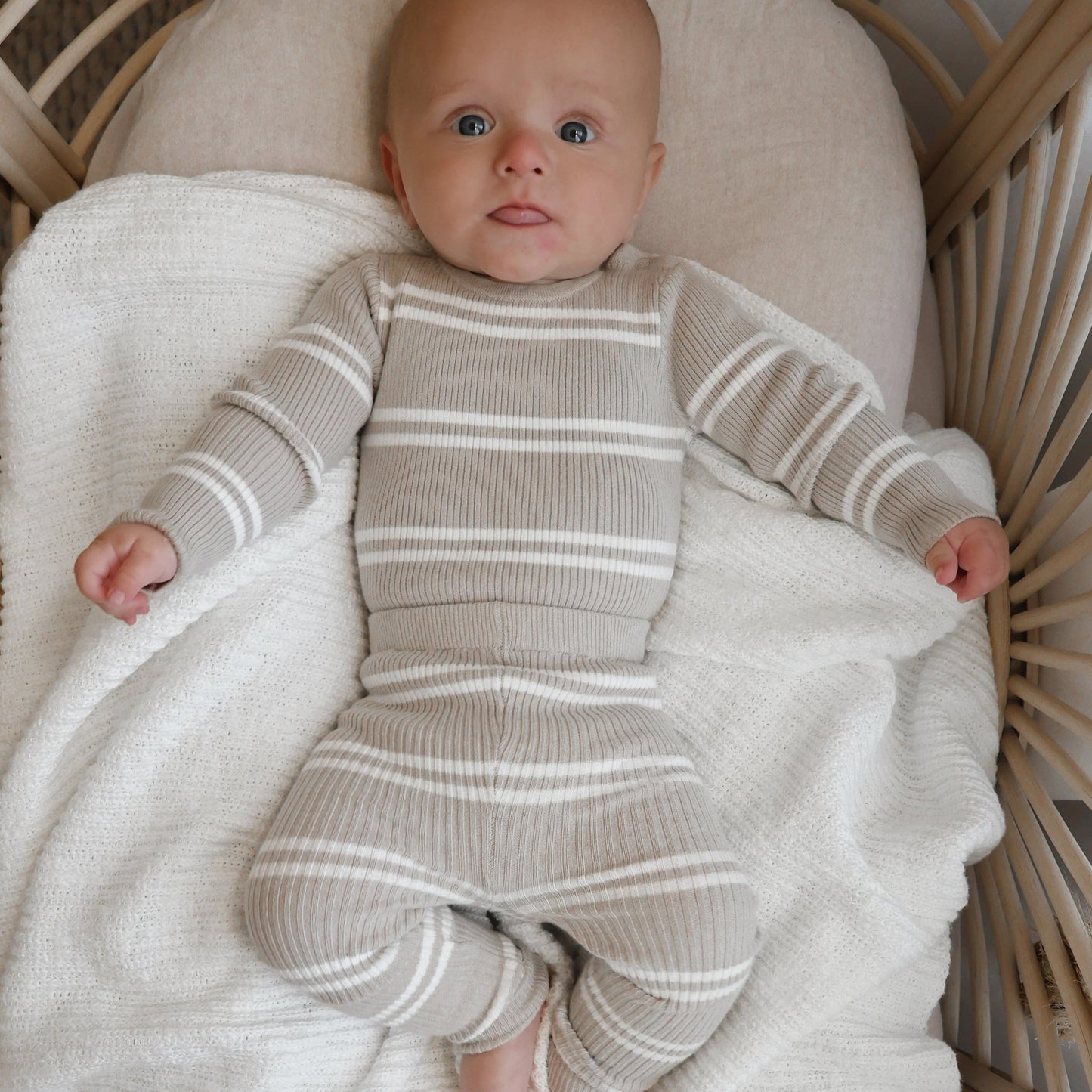 Baby in a striped outfit lying on a white blanket in a wicker crib.