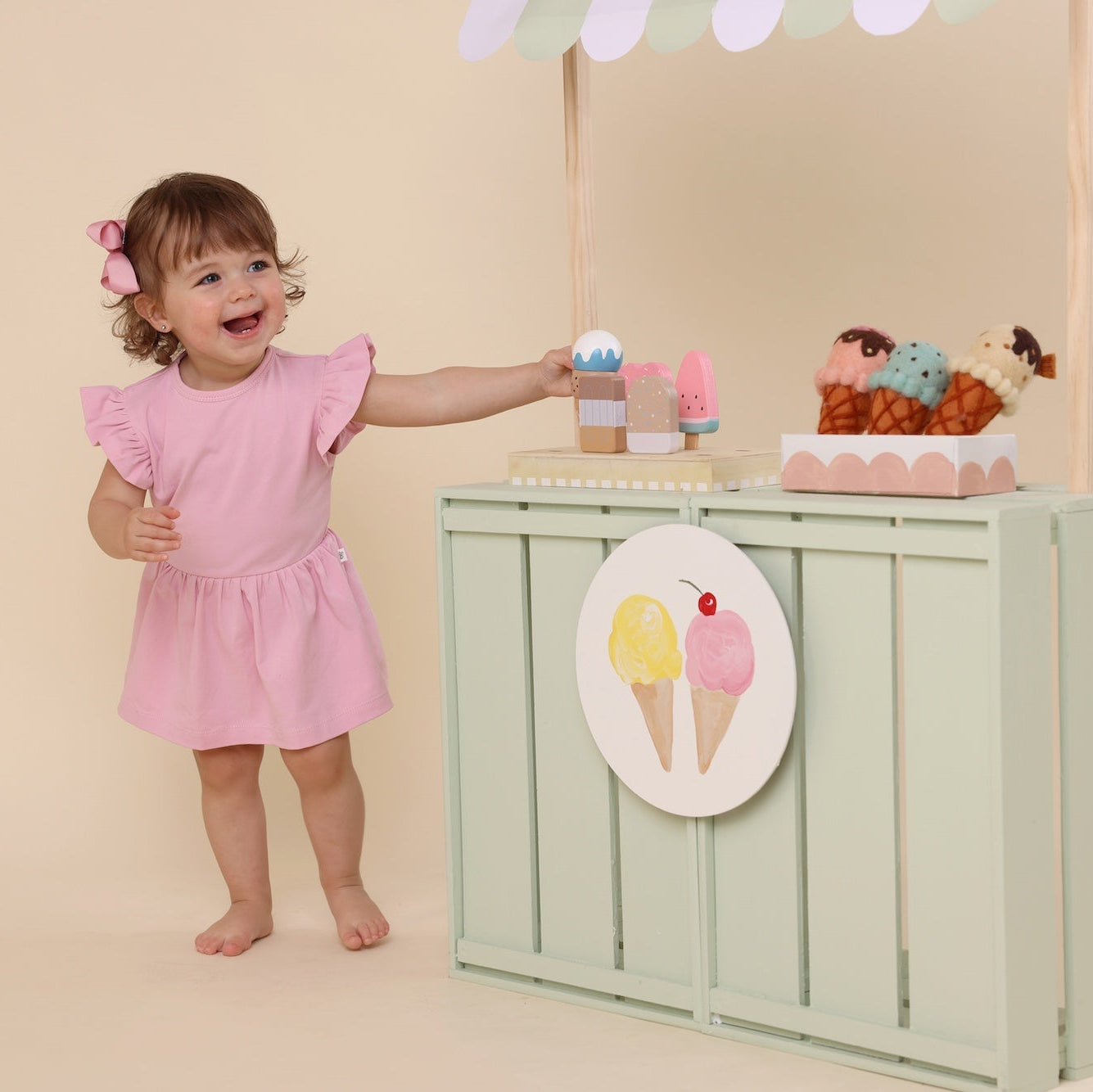 Child in a pink dress standing next to a toy ice cream stand with various toys on a beige background