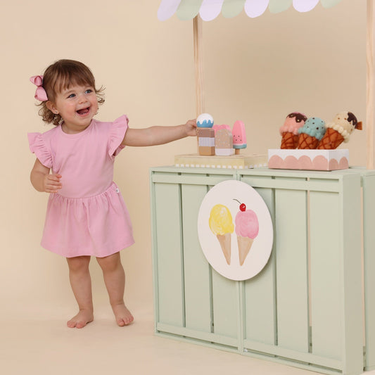 Child in a pink dress standing next to a toy ice cream stand with various toys on a beige background