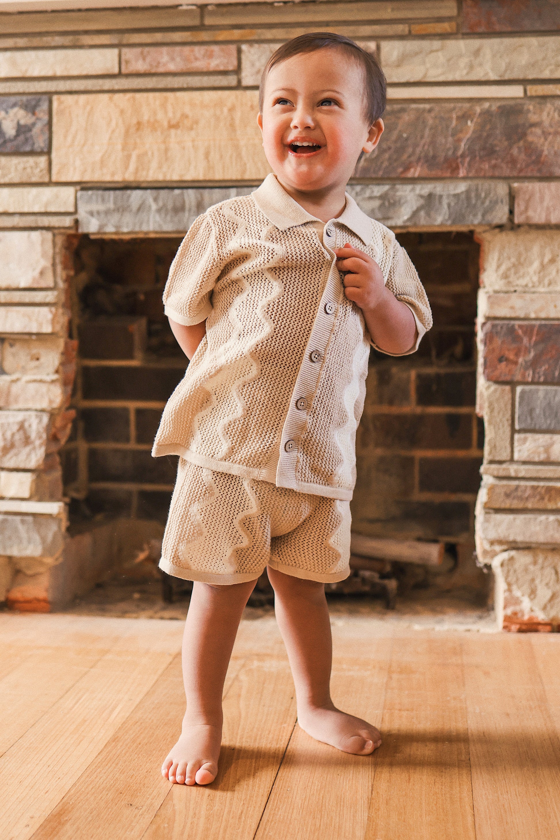 Child wearing a beige outfit standing in front of a stone fireplace.