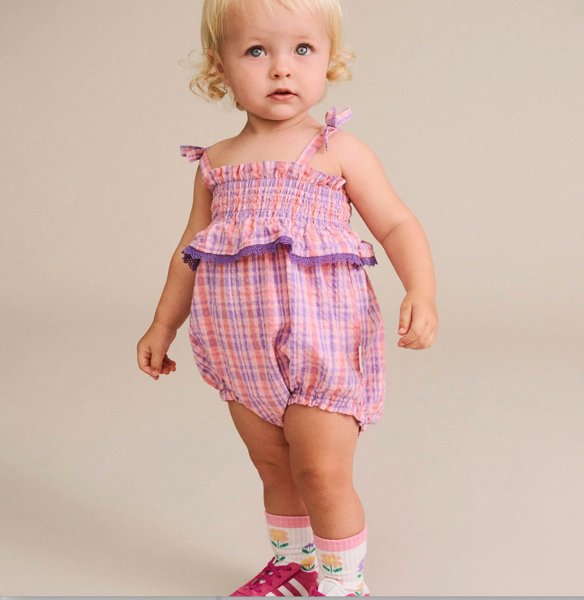 A toddler girl wearing a pink and purple plaid romper with ruffled trim, standing against a plain beige studio background.