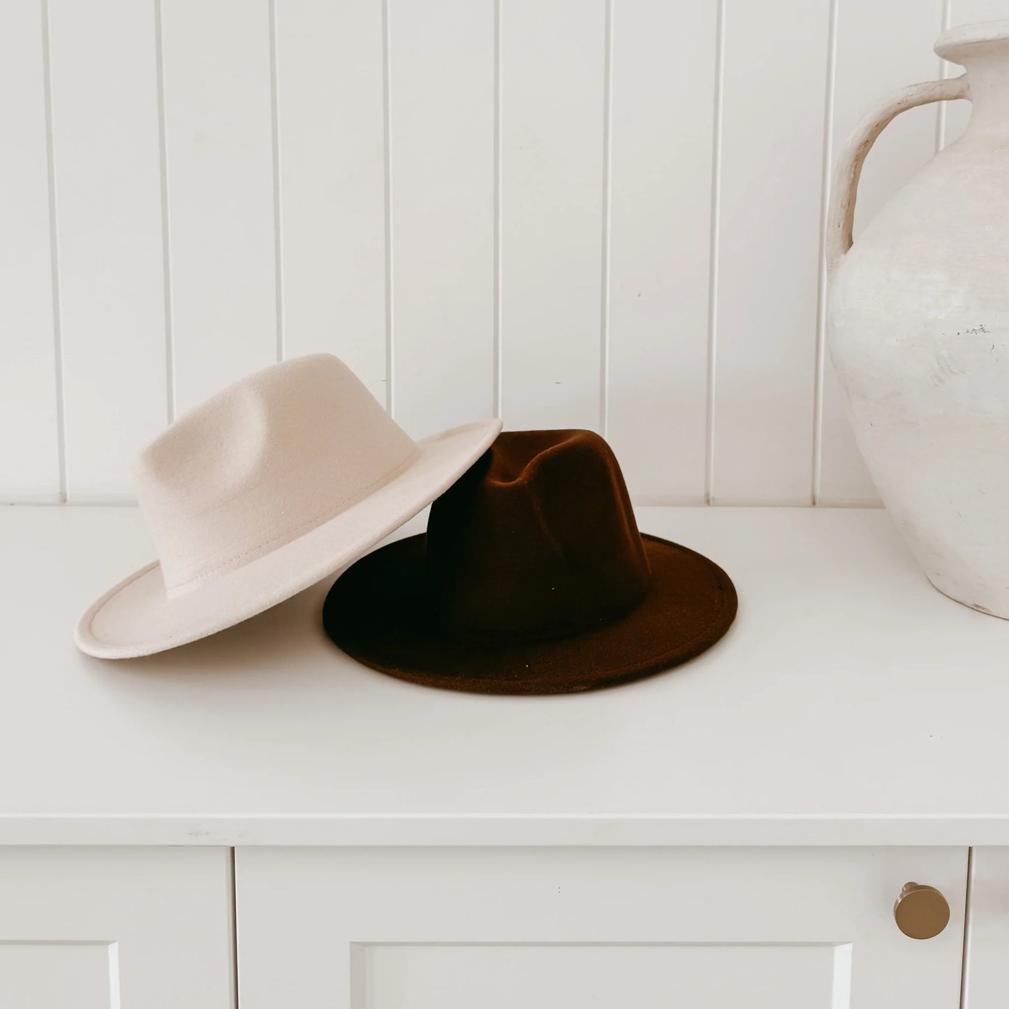 Two hats, one beige and one brown, on a white surface with a white wooden panel background.