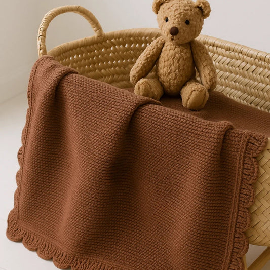 Brown teddy bear sitting on a brown knitted blanket in a woven basket.