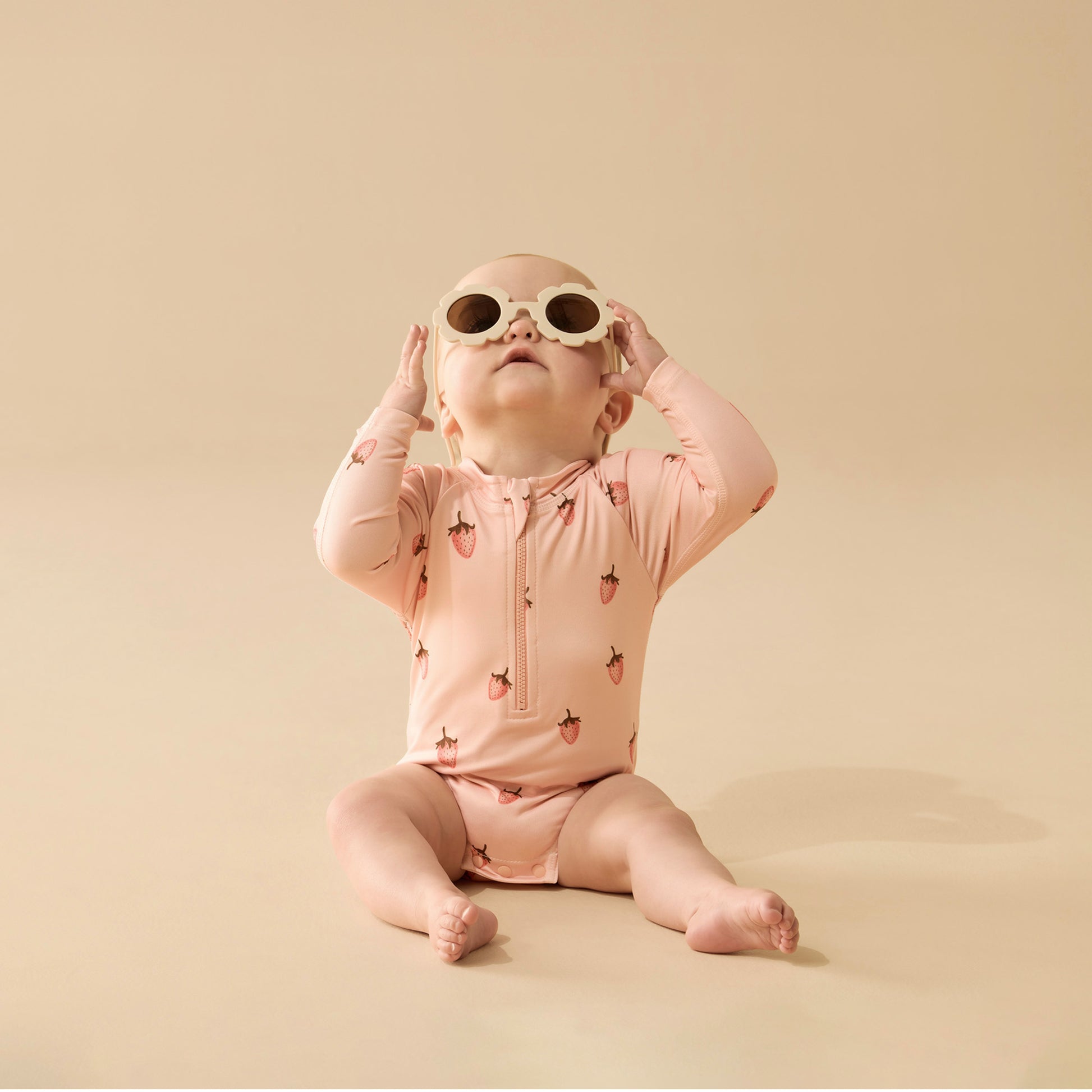 Baby wearing a pink strawberry-patterned onesie and sunglasses on a beige background