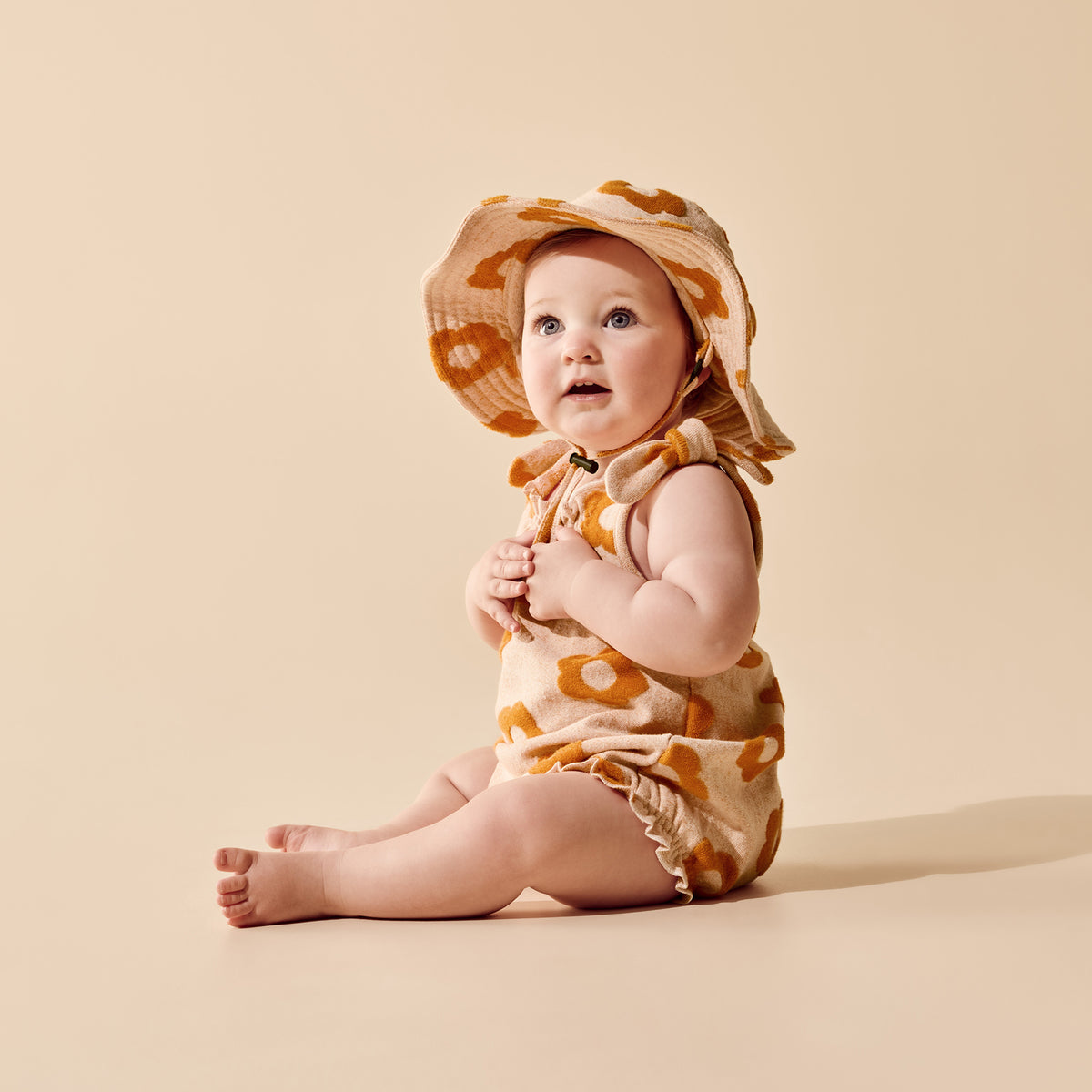A baby sitting on a warm beige studio background, wearing a sleeveless cream romper and matching wide-brim sun hat with a mustard yellow abstract pattern.