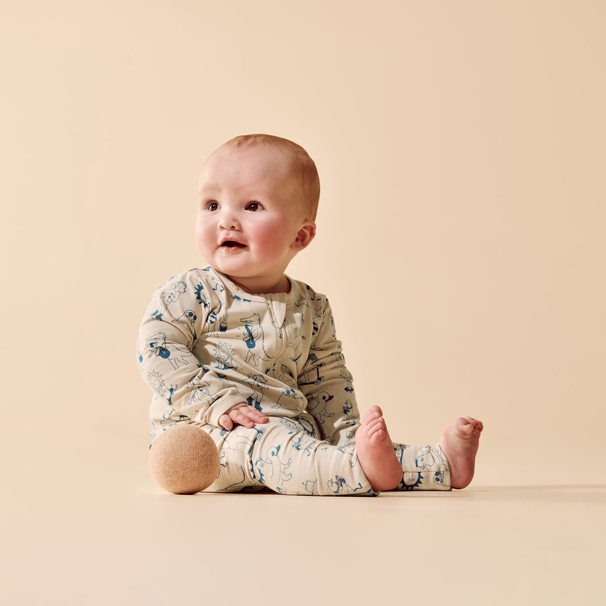 A baby sitting on a warm beige studio background, wearing a long-sleeve cream-colored romper with a blue and brown animal print.