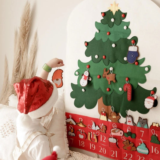 A child wearing a red Santa hat sits on a cream knit blanket, interacting with a large felt Christmas tree advent calendar mounted on a wall. The red advent section at the bottom shows numbered pockets with festive illustrations.