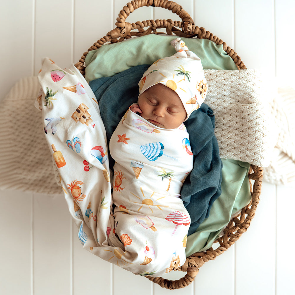 Newborn baby swaddled in colorful blankets with a headband, lying in a woven basket against a white wooden panel background.
