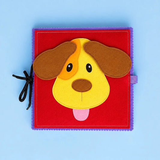 A square felt baby activity book or soft toy with a colorful dog face design, featuring a red center, purple border, brown floppy ears, a yellow face, black eyes, and a pink tongue, photographed on a light blue background.