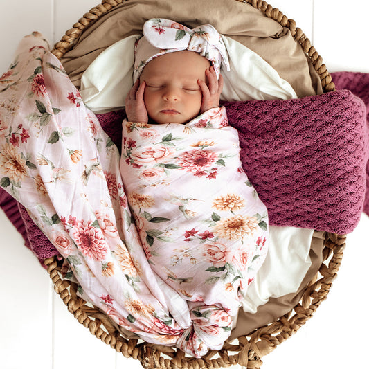 Newborn baby wrapped in a floral blanket with a matching headband and knitted blanket in a wicker basket.