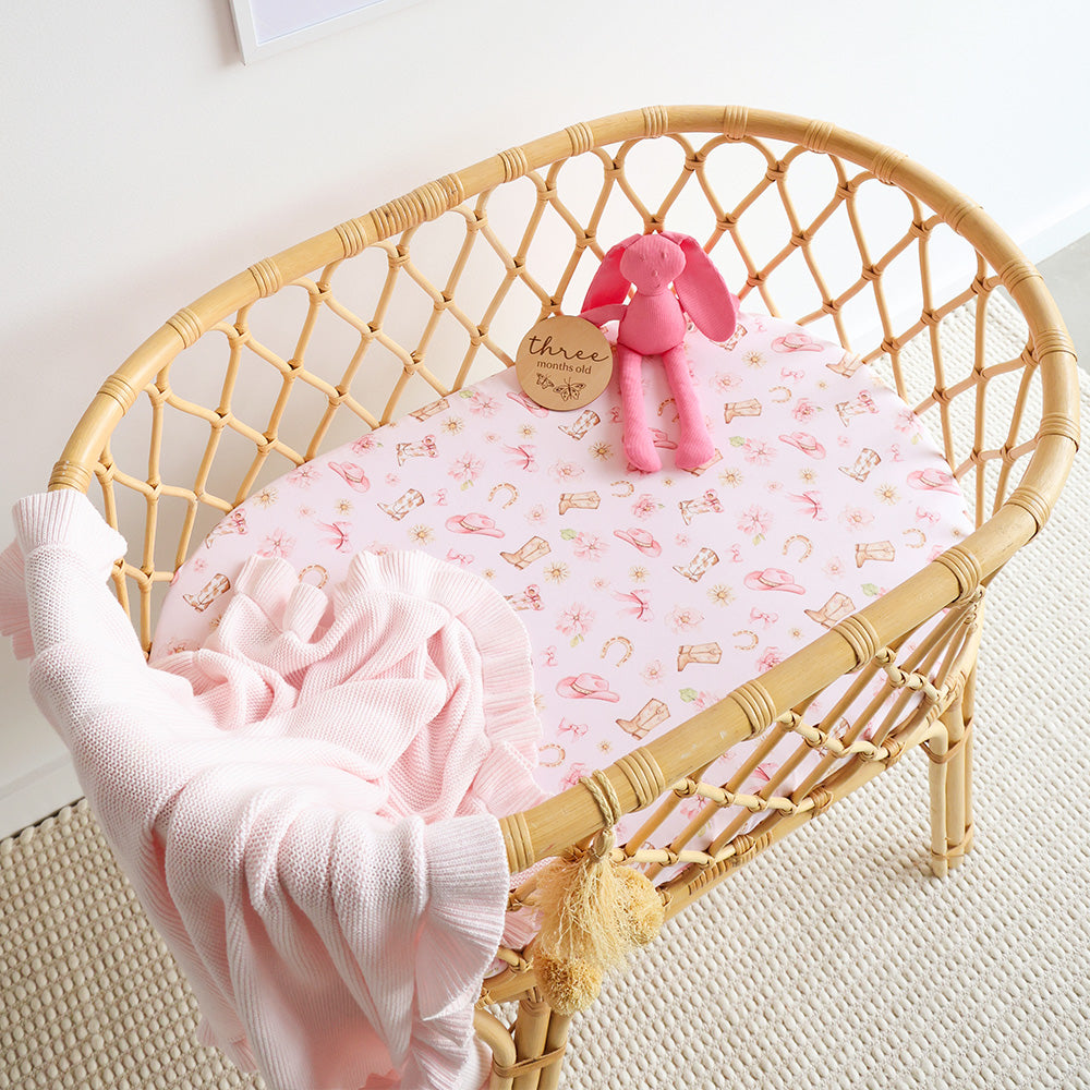 A light natural rattan baby bassinet styled with a pink patterned mattress, a pink plush bunny toy, and a soft pink blanket draped over the side, set on a textured cream rug.