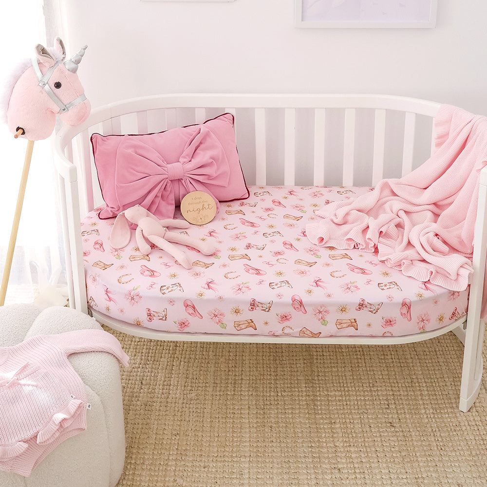 A pink fitted crib sheet with a repeating unicorn print is shown in a white crib, styled with a pink bow pillow, plush bunny, blanket, and a unicorn toy in a bright nursery setting.