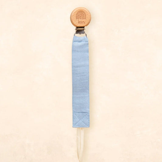 A light blue fabric strap accessory with a round natural wood end and a small metal connector, shown against a warm beige textured background.