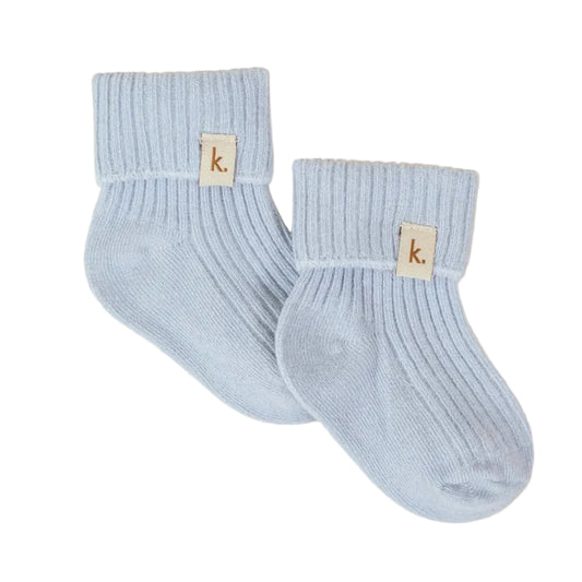 A pair of light blue ribbed ankle socks with a small beige brand tag featuring a lowercase 'k' logo, displayed on a textured cream background.