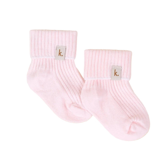 A pair of light pink ribbed ankle socks with a small square gray brand patch featuring the letter 'k' on the cuff, displayed on a textured beige background.