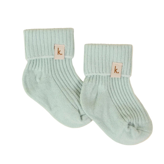 A pair of light mint green ribbed knit baby socks with a small beige leather patch featuring the lowercase 'k' logo, displayed on a textured cream background.