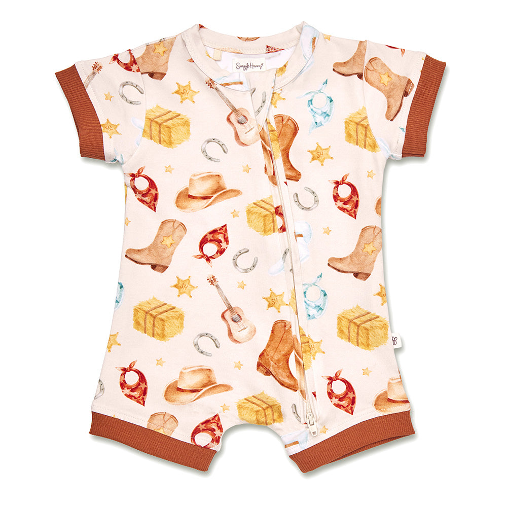 A baby short-sleeve romper with a western-themed print featuring cowboy boots, hats, bandanas, guitars, and stacked boxes on a cream background, finished with brown ribbed trim.