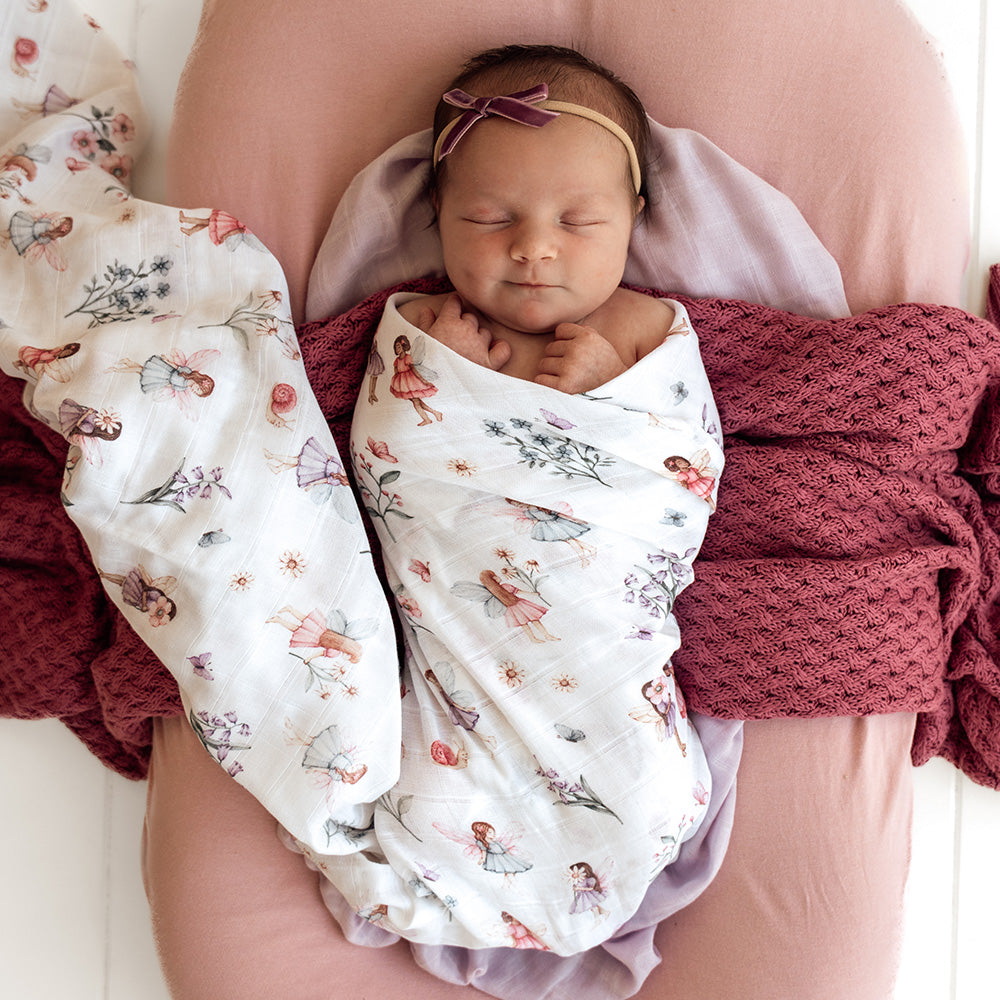 Newborn baby swaddled in a floral blanket with pink headband, lying on a pink surface.