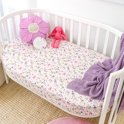 A white wooden crib with a colorful floral-patterned mattress cover, styled with a pink flower pillow, a pink plush bunny, a round tag, and a purple knit blanket.