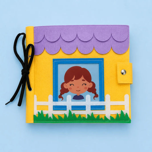 A handmade felt journal or book cover featuring a house design with a purple scalloped roof, yellow body, white picket fence, green grass, and a cartoon girl in a blue window frame. A black ribbon is tied to the left side, and the item is photographed on a light blue background.
