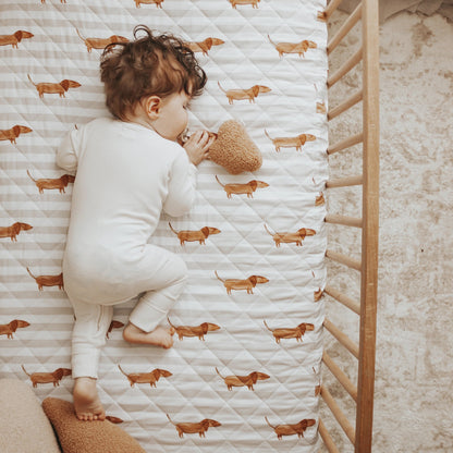 Child lying on a crib mattress with dachshund pattern, surrounded by wooden crib and pillows.