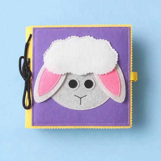 A square purple felt baby activity book featuring a sheep face applique with white wool, pink inner ears, and a gray face. The book has a yellow stitched border, a black hanging loop with a black cord, and a yellow tab on the side, set against a solid light blue background.