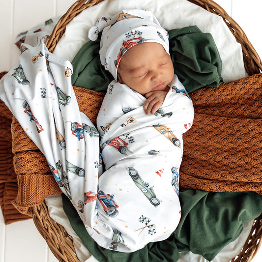 Newborn baby swaddled in a car-themed blanket in a wicker basket with brown and green blankets.