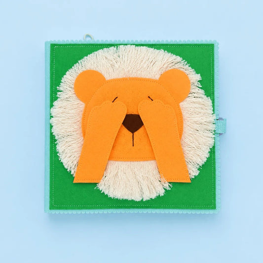 A square fabric sensory toy featuring a lion face design, with orange felt ears and face, cream-colored faux fur mane, and a green background, shown on a light blue backdrop.
