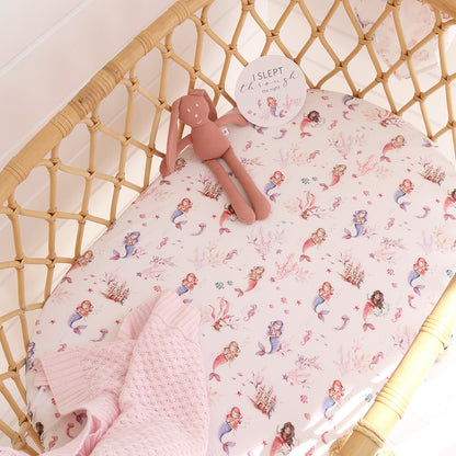 Baby crib with mermaid-patterned sheet, pink blanket, and plush toy.