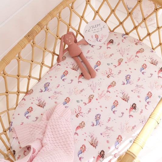 Baby crib with mermaid-patterned sheet, pink blanket, and plush toy.