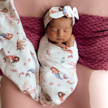 Newborn baby wrapped in a mermaid-themed swaddle with a matching headband, lying on a pink surface.