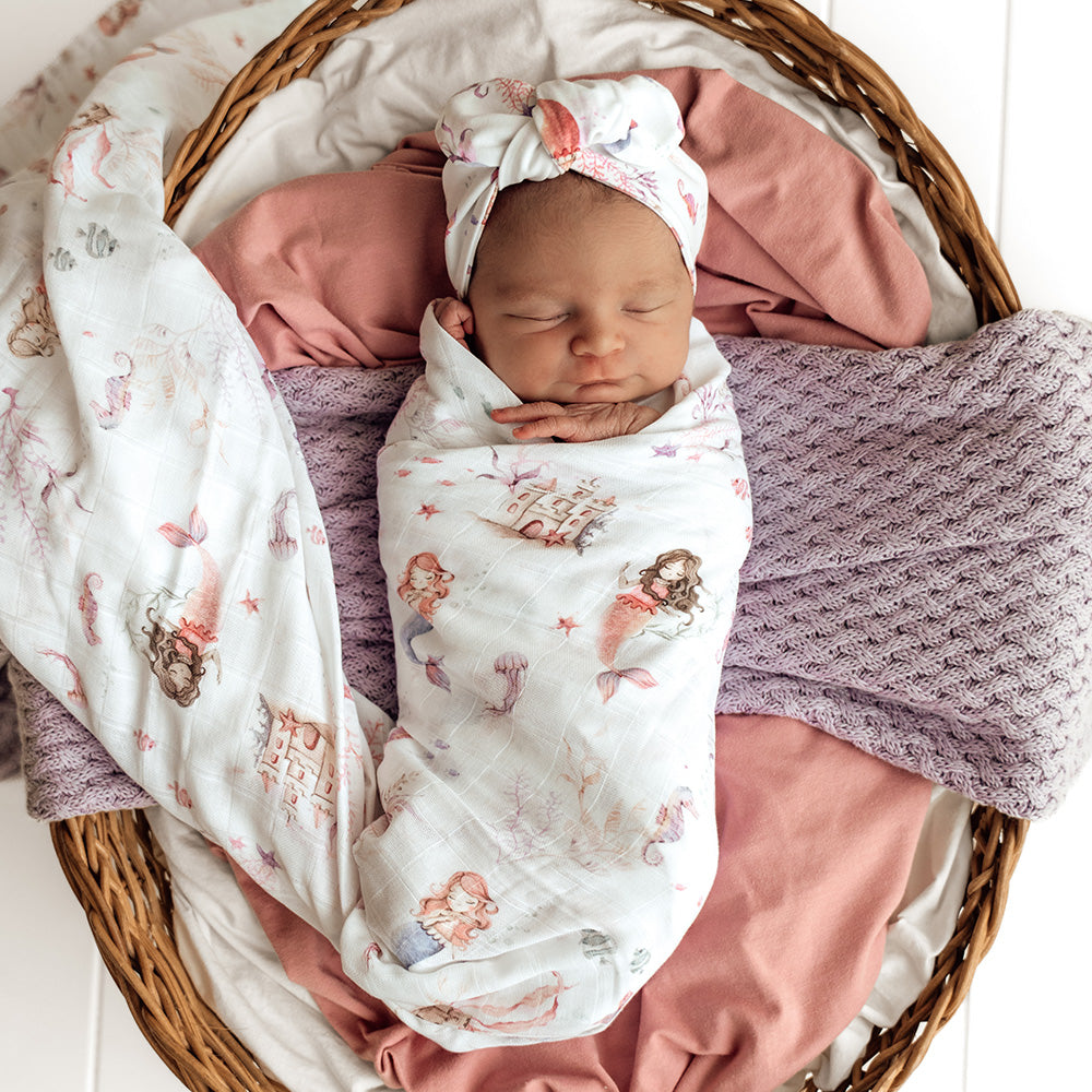 Newborn baby wrapped in a floral blanket with pink pants and a matching headband, lying in a wicker basket.