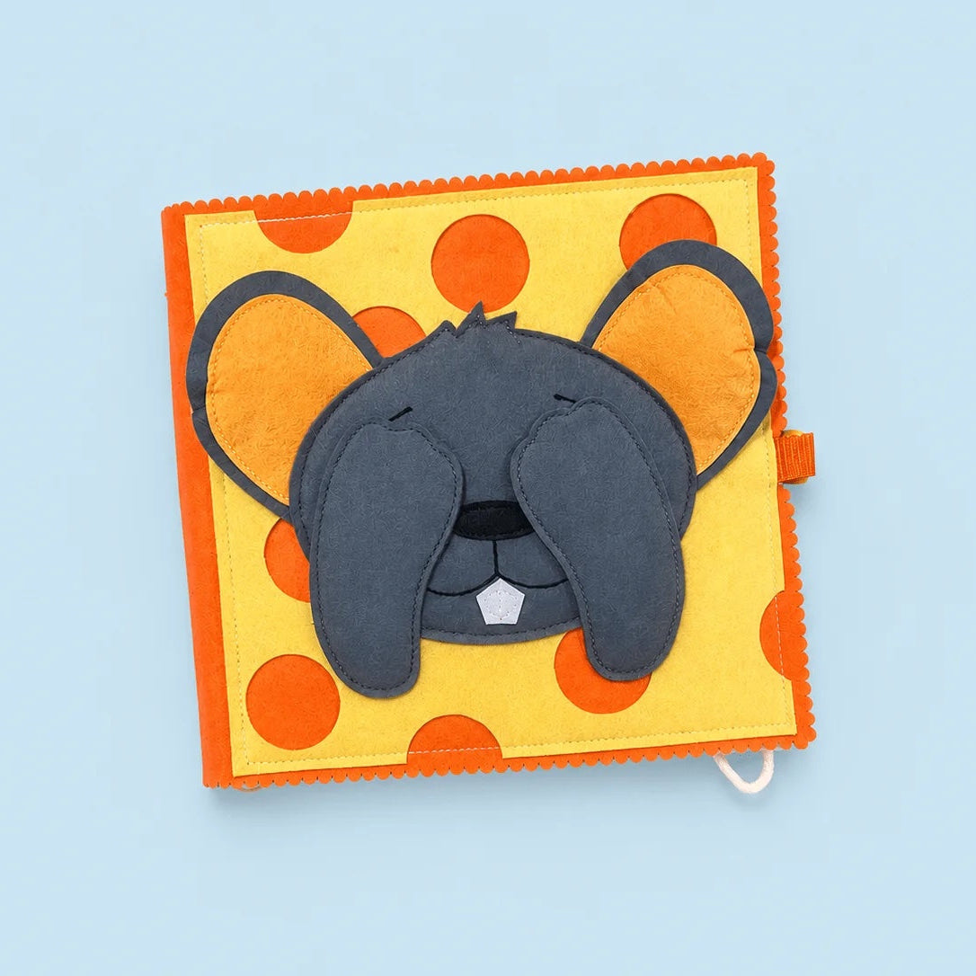 A square fabric sensory toy featuring a gray mouse face with closed eyes on a yellow background with orange polka dots, set against a light blue background.