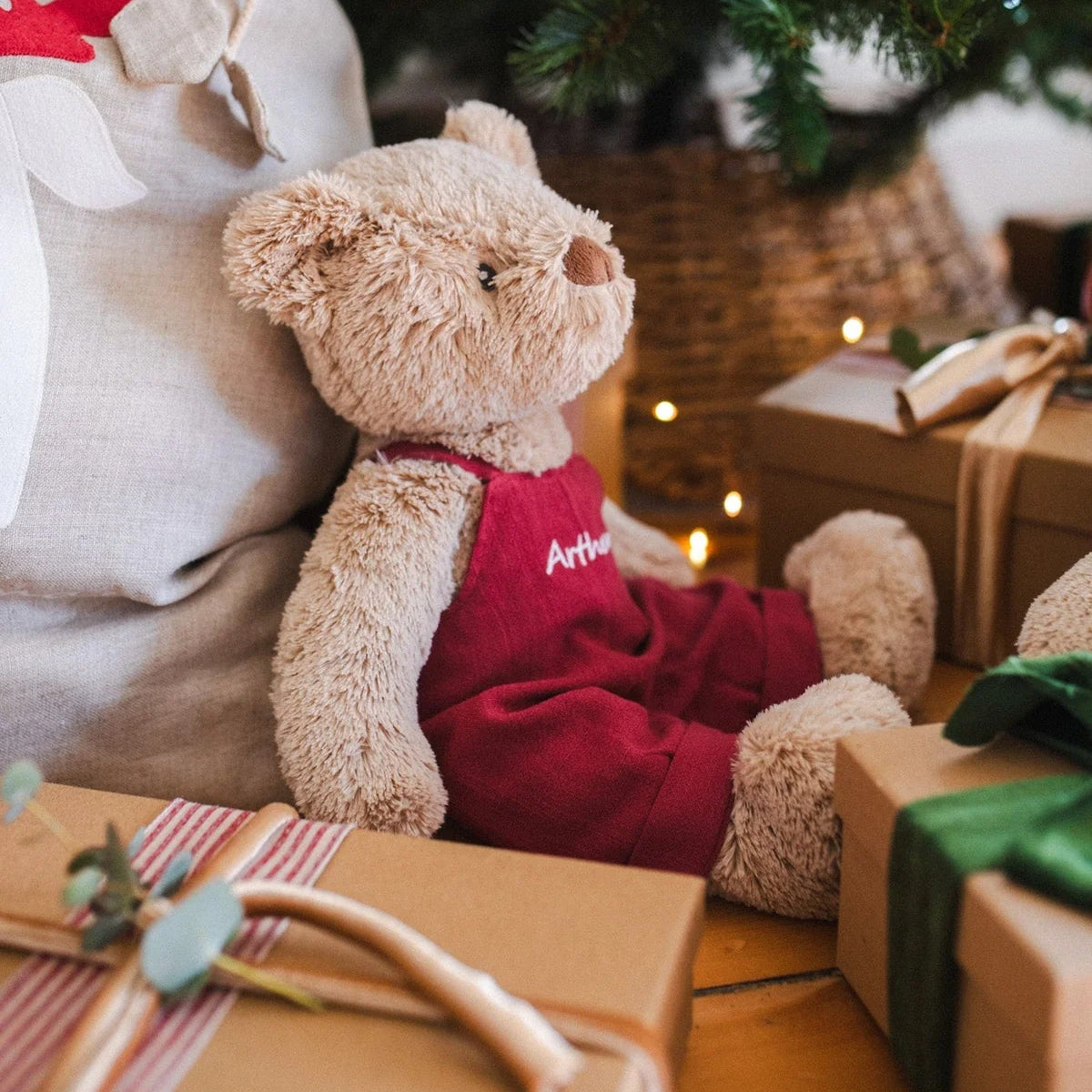 Teddy bear in a red outfit sitting under a decorated Christmas tree with presents around.