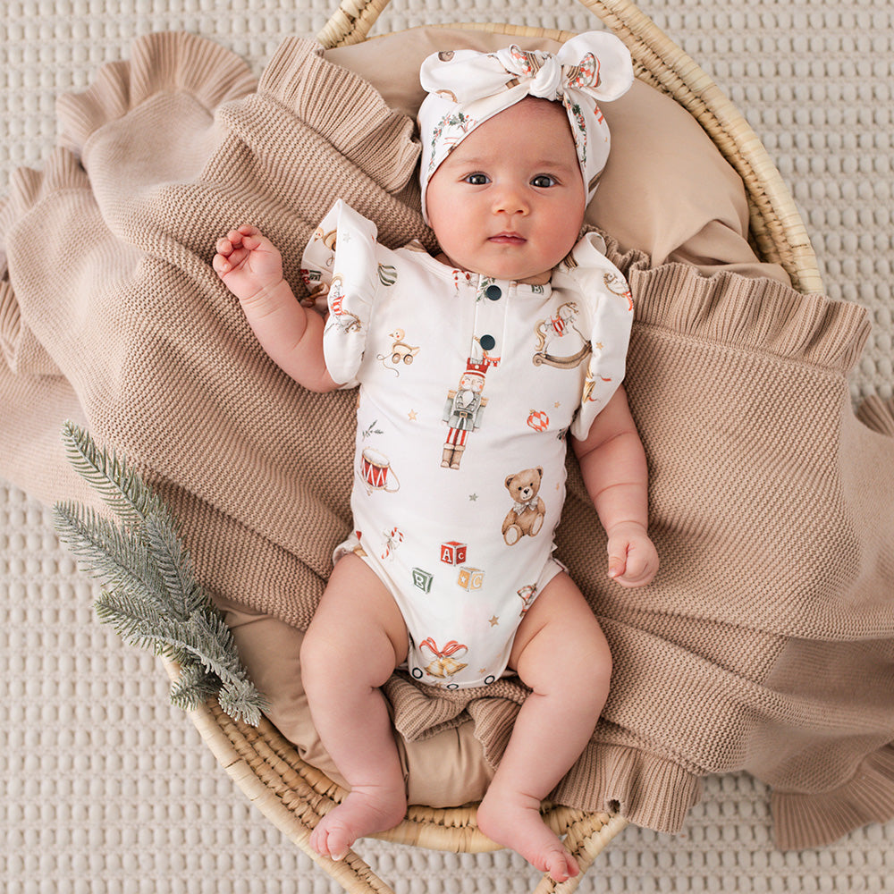 A baby lying on a woven basket with a beige blanket, wearing a white short-sleeve bodysuit with a teddy bear and toy print, plus a matching white bow headband.