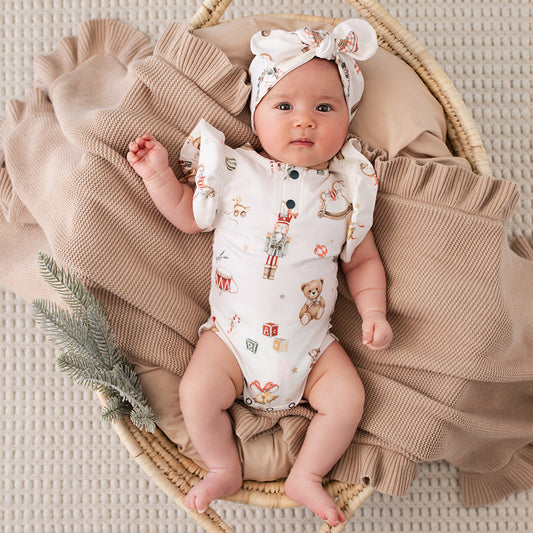 A baby lying on a woven basket with a beige blanket, wearing a white short-sleeve bodysuit with a teddy bear and toy print, plus a matching white bow headband.